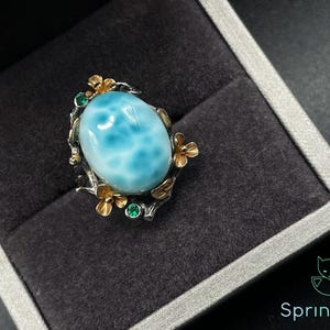 Könnte beinhalten: Ein silberner Ring mit einem großen, ovalen, hellblauen Edelstein. Der Ring ist mit goldfarbenen floralen Details und kleinen grünen Edelsteinen verziert. Der Ring wird in einer schwarzen Schmuckschatulle präsentiert.