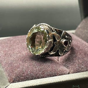 Könnte beinhalten: Ein Silberring mit einem großen, facettierten, hellgrünen Edelstein. Der Ring hat aufwendige florale und seilartige Details, mit einem kleinen, runden, roten Stein. Der Ring wird auf einer violetten Samtoberfläche präsentiert.