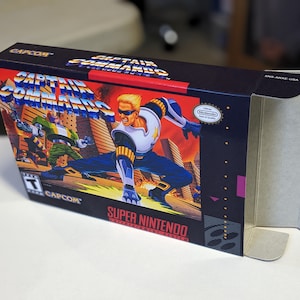 May include: A Super Nintendo game cartridge box for Captain Commando. The box is black with a red and yellow design. The front of the box features a character in a blue and white suit with a yellow star on his chest. The box is labeled "CAPCOM" and "SUPER NINTENDO" in white text.