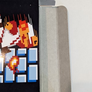 Pu&ograve; includere: Una scatola di cartone con un personaggio di videogioco sul davanti. Il personaggio &egrave; in stile pixelato e indossa un abito rosso e bianco. Il personaggio sta saltando e ci sono delle fiamme dietro di lui. La scatola &egrave; piegata e i bordi sono visibili.