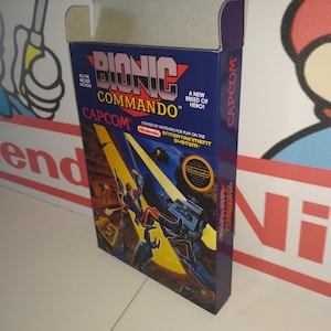 May include: A blue and yellow box for the Nintendo Entertainment System video game Bionic Commando. The box features a character in a red and blue suit holding a gun and fighting a robot. The box has the text "Bionic Commando" and "Capcom" on it.