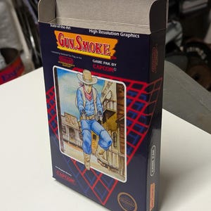 May include: A vintage Gun Smoke video game box. The box is dark blue with a cowboy illustration. Text includes "Gun Smoke", "Nintendo Entertainment System", and "CAPCOM". The box is rectangular and has an open top.