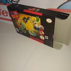 Puede incluir: Una caja de juego de Super Nintendo para Earthworm Jim 2. La caja es negra con un diseño amarillo y rojo. La parte delantera de la caja presenta un personaje de dibujos animados con un traje verde saltando. El texto "Earthworm Jim 2" está impreso en rojo y blanco en la caja.