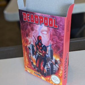 May include: A red and black video game cartridge box with the title "DEADPOOL" in large red letters. The box features an illustration of Deadpool in a red and black suit, holding two katanas, standing in front of a cityscape with flames and smoke. The box also includes the text "Official Nintendo Seal" in a gold circle.