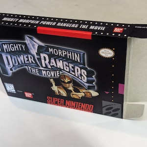 May include: A black Super Nintendo game cartridge box with a red stripe. The box features a white and silver logo for "Mighty Morphin Power Rangers: The Movie". The front of the box shows a white Power Ranger with a tiger helmet, holding a sword.