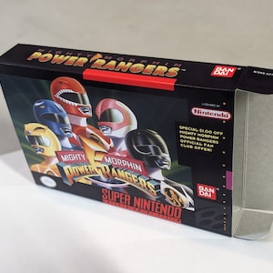 May include: A black Super Nintendo game cartridge box with a red, white, and blue Mighty Morphin Power Rangers logo. The box features a picture of the Power Rangers in their colorful suits. The text "SPECIAL $1.00 OFF MIGHTY MORPHIN POWER RANGERS OFFICIAL FAN CLUB OFFER!" is printed on the box.