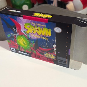 Puede incluir: Una caja de videojuego de Super Nintendo para "Spawn". La caja presenta una ilustración vibrante del personaje de Spawn con una capa roja, con el título del juego en amarillo y el texto "Todd McFarlane's Spawn: The Video Game".