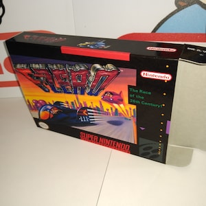 May include: A Super Nintendo game box for the game "F-Zero: The Race of the 26th Century!" The box is black with a red and yellow image of a futuristic race car on the front. The game is a racing game.