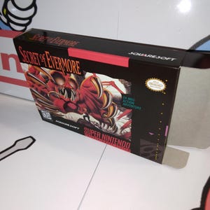 May include: A black video game box for "Secret of Evermore" on Super Nintendo. The box features a red and white monster illustration. The text "SQUARESOFT" and "SUPER NINTENDO" are visible.