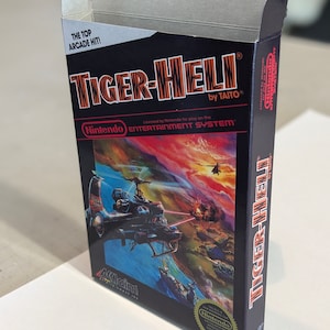 Può includere: Una scatola di cartuccia di gioco vintage per Nintendo Entertainment System per Tiger-Heli di Taito. La scatola presenta un'illustrazione colorata di un elicottero che vola sopra un paesaggio con esplosioni e altri veicoli militari. Il testo "THE TOP ARCADE HIT!" è stampato sulla parte superiore della scatola.