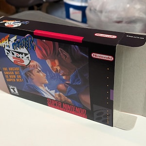 Może przedstawiać: Pudełko z grą Super Nintendo dla Street Fighter Alpha 2. Pudełko jest czarne z czerwonym akcentem i zawiera kolorową ilustrację dwóch postaci w pozycji bojowej. Widoczny jest tekst "Street Fighter Alpha 2" i "Super Nintendo".
