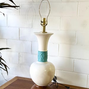 Może przedstawiać: Wintage lampa stołowa z kremowym ceramicznym korpusem, turkusowym pasem z kwiatowym wzorem i ciemnobrązową drewnianą podstawą. Lampa ma mosiężną szyję i brązowe zwieńczenie. Lampa jest podłączona.