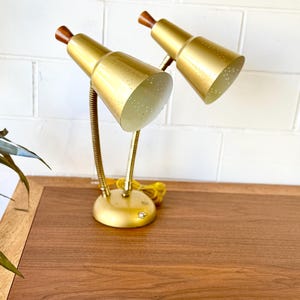 Puede incluir: Un par de lámparas de escritorio doradas vintage con brazos de cuello de cisne ajustables. Cada lámpara presenta una pantalla cónica con pequeñas perforaciones y un acento de madera. Las lámparas están sobre una base redonda y sobre una superficie de madera.