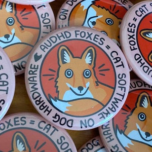 May include: A cluster of pink circular badges featuring a cartoon fox design. The fox is depicted in orange and white, encircled by text that reads "AUDHD FOXES = CAT SOFTWARE ON DOG HARDWARE NO".