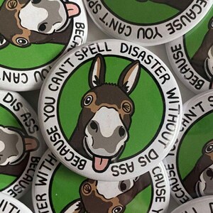 May include: A pile of white, circular badges featuring a cartoon donkey with a brown and white face. The text on the badge reads, "BECAUSE YOU CAN'T SPELL DISASTER WITHOUT DIS ASS." The donkey has its tongue sticking out.