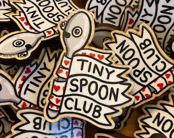 May include: Wooden Tiny Spoon Club pins. Each pin features a cartoon spoon holding a banner with the words "Tiny Spoon Club." The banner is white with black text and red heart accents. The pins are made of wood with a natural finish.