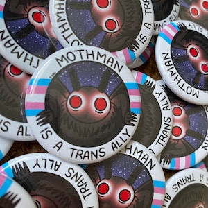 May include: A pile of circular badges displaying a cartoon Mothman with red eyes against a starry backdrop. The badges read "Mothman is a Trans Ally" and incorporate the trans pride flag colours.