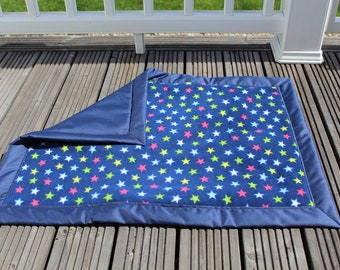 Può includere: Una coperta in pile blu con un bordo blu scuro e un motivo di stelle colorate. La coperta è piegata a metà e si trova su un ponte di legno.