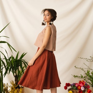 May include: A woman wearing a light brown sleeveless top and a rust-colored skirt. She is standing in front of a white backdrop with plants and flowers.