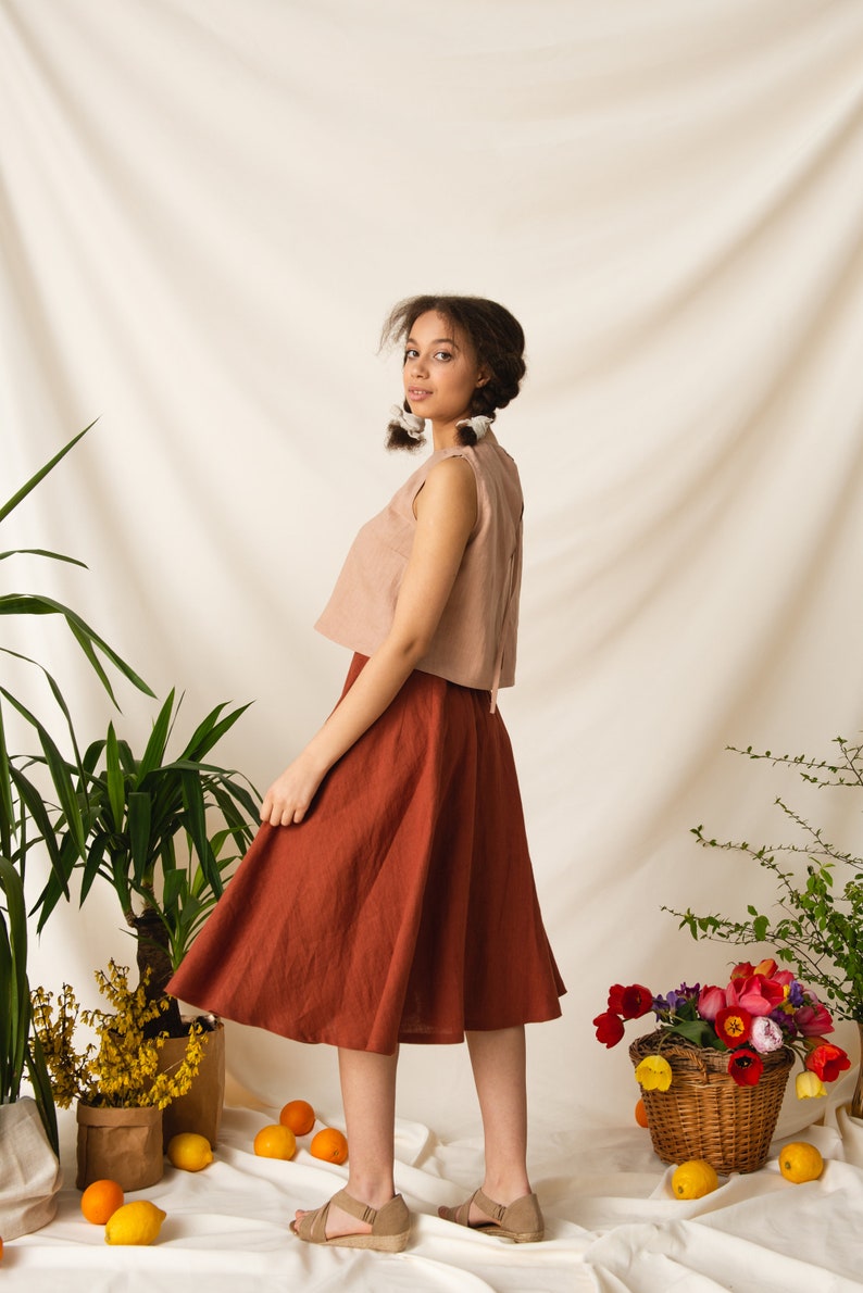 May include: A woman wearing a light brown sleeveless top and a rust-colored skirt. She is standing in front of a white backdrop with plants and flowers.