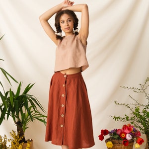 May include: A woman wearing a light brown linen top and a rust-colored linen skirt with buttons down the front. She is standing in front of a white backdrop with plants and lemons.