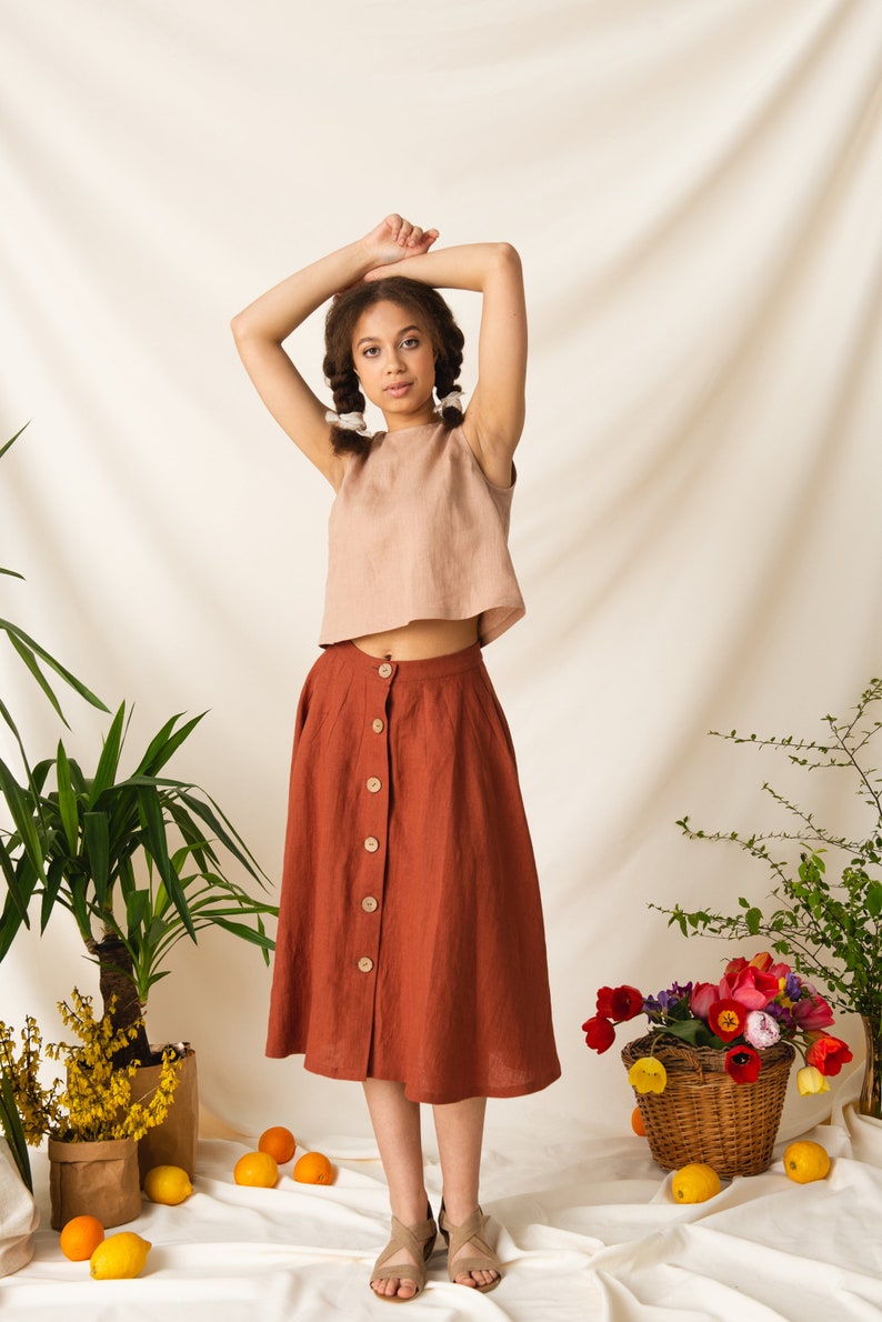 May include: A woman wearing a light brown linen top and a rust-colored linen skirt with buttons down the front. She is standing in front of a white backdrop with plants and lemons.