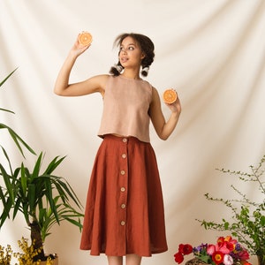 May include: A woman wearing a light brown sleeveless top and a rust-colored skirt with buttons down the front. She is holding two orange slices in her hands. The background is a white backdrop with plants and flowers.