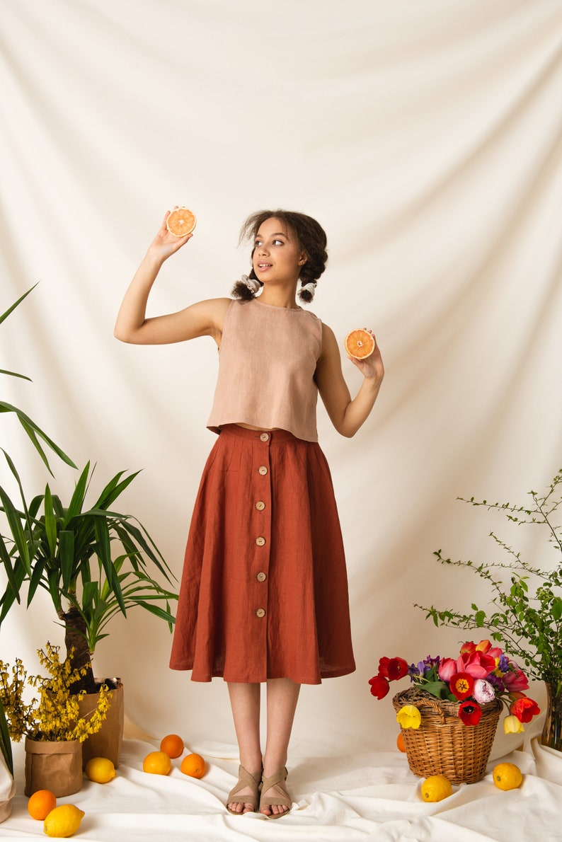 May include: A woman wearing a light brown sleeveless top and a rust-colored skirt with buttons down the front. She is holding two orange slices in her hands. The background is a white backdrop with plants and flowers.