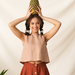 May include: A young person with brown hair in braids is wearing a light brown sleeveless top and a rust-colored skirt with buttons down the front. They are holding a pineapple above their head and smiling at the camera.