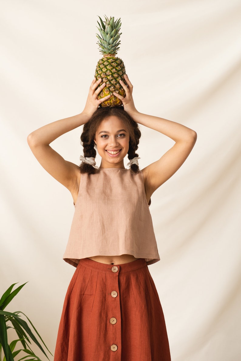 May include: A young person with brown hair in braids is wearing a light brown sleeveless top and a rust-colored skirt with buttons down the front. They are holding a pineapple above their head and smiling at the camera.
