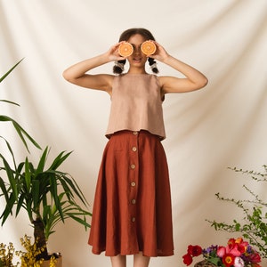 May include: A woman wearing a light brown sleeveless top and a rust-colored skirt with buttons down the front. She is holding two halves of an orange in front of her eyes. The background is a white fabric backdrop with plants and flowers.