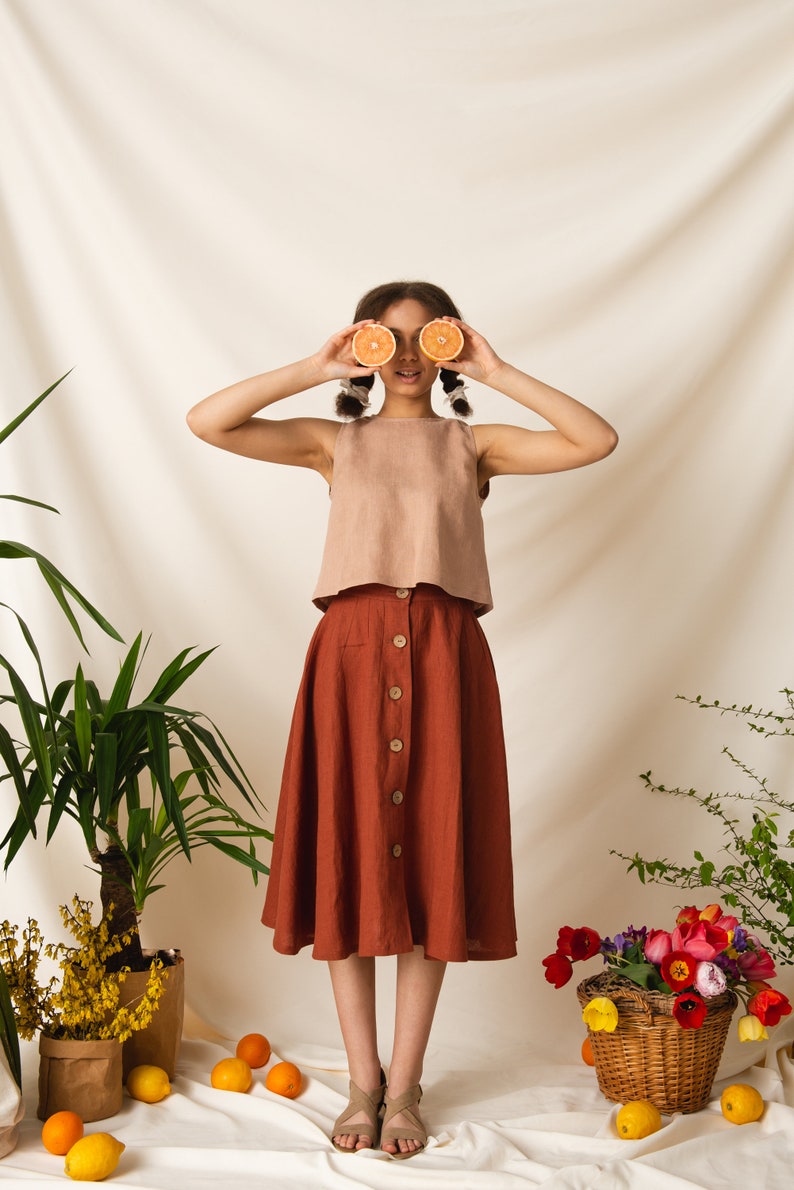May include: A woman wearing a light brown sleeveless top and a rust-colored skirt with buttons down the front. She is holding two halves of an orange in front of her eyes. The background is a white fabric backdrop with plants and flowers.