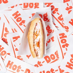 Puede incluir: Un perrito caliente recién hecho en un bollo, relleno de ensalada de col blanca y naranja, descansa sobre un lecho de bolsas de papel blancas para perritos calientes con texto rojo. Las bolsas llevan la inscripción "Hot-Dog".