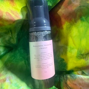 May include: A clear bottle of "Peppermint 'Kwoodchic' Cleanser" with a dark blue cap. The label is pink and white, with product information and ingredients. The bottle is set against a colorful, tie-dye background of green, yellow, and orange.