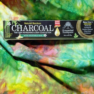 Op de afbeelding: Een zwarte doos met Natural Bamboo Charcoal tandpasta met witte tekst en accenten. De doos leest "Essential Toothpaste" en "Netto gewicht 184g." De achtergrond is een kleurrijke tie-dye stof met groene, gele en oranje tinten.