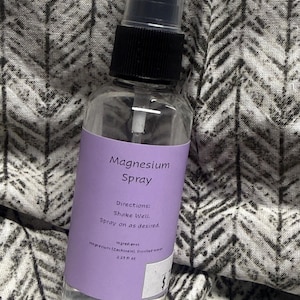 May include: A clear plastic spray bottle with a black pump top and a light purple label. The label reads Magnesium Spray, Directions: Shake Well. Spray on as desired. Ingredients: Magnesium (Zechstein), Distilled water. 2.25 fl oz.