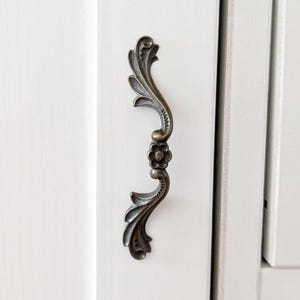 Peut inclure: Une poignée de placard verticale avec une finition bronze antique, présentant un design orné et incurvé avec des motifs de feuilles et une fleur centrale. La poignée est fixée sur une surface verticale blanche avec une jointure visible.