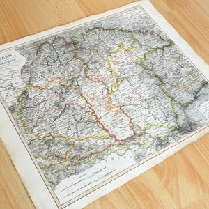 Puede incluir: Un mapa de papel antiguo titulado Ungarn und das Theil von Siebenbürgen, que presenta líneas topográficas detalladas, fronteras regionales en rojo, amarillo y verde, y etiquetas de texto impresas. El mapa incluye una escala.