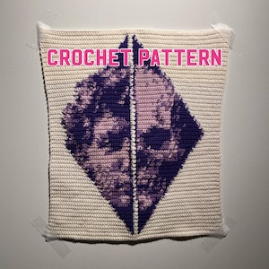 Può includere: Uno schema di uncinetto bianco con una forma di diamante viola al centro. La forma di diamante presenta un disegno di due volti, uno su ogni lato. Il testo "CROCHET PATTERN" è scritto in rosa nella parte superiore dello schema.