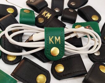 May include: A close-up of green and black leather cord organizers with gold metal accents. The organizers have the initials "KM" embossed in gold on the green leather.