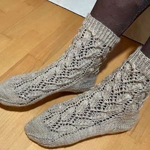 Könnte beinhalten: Ein Paar beige gestrickte Socken mit einem zarten Spitzenmuster. Die Socken sind aus feinem Garn gefertigt und haben ein weiches, angenehmes Tragegefühl.
