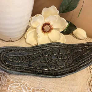 Könnte beinhalten: Eine dunkelgraue Keramikschale mit einem erhabenen Blumendesign. Die Schale ist oval geformt und hat einen leicht abgerundeten Rand.