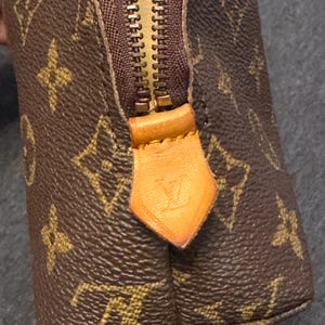 May include: Brown and tan monogrammed cosmetic bag with a gold zipper and leather pull tab. The bag features a repeating pattern of interlocking letters and floral designs. The zipper pull has a debossed logo.