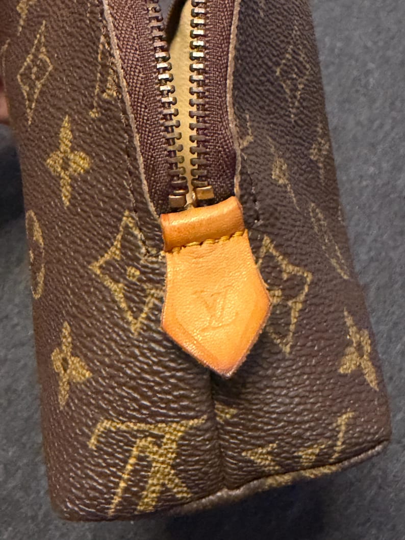 May include: Brown and tan monogrammed cosmetic bag with a gold zipper and leather pull tab. The bag features a repeating pattern of interlocking letters and floral designs. The zipper pull has a debossed logo.