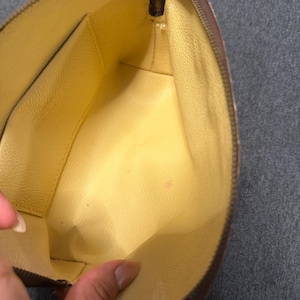 May include: Open view of a brown zippered pouch with a yellow interior. The pouch has a rectangular shape with rounded edges. The zipper is gold-colored. The interior has a small pocket on one side.