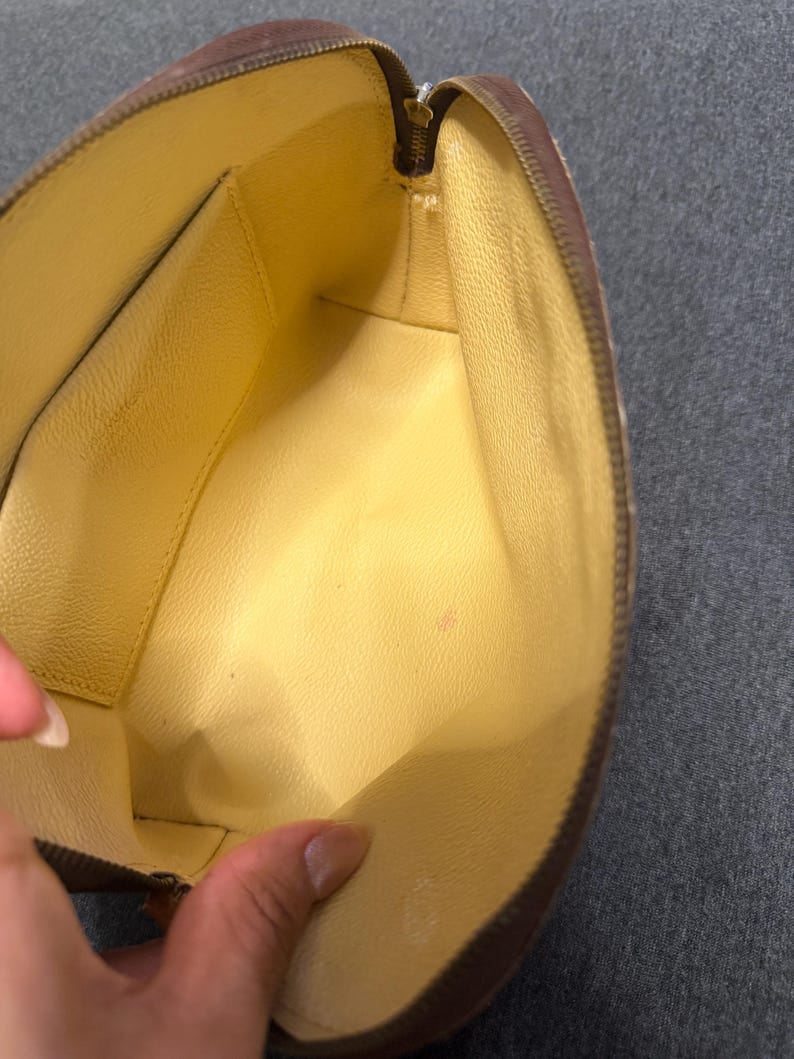 May include: Open view of a brown zippered pouch with a yellow interior. The pouch has a rectangular shape with rounded edges. The zipper is gold-colored. The interior has a small pocket on one side.