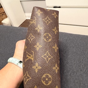 May include: Brown and gold monogrammed clutch. The clutch features a repeating pattern of interlocking initials and floral motifs. The bag is held in a person's hand, with a light blue and green bracelet visible on the wrist.