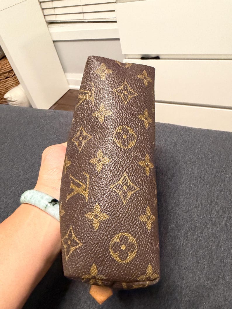 May include: Brown and gold monogrammed clutch. The clutch features a repeating pattern of interlocking initials and floral motifs. The bag is held in a person's hand, with a light blue and green bracelet visible on the wrist.