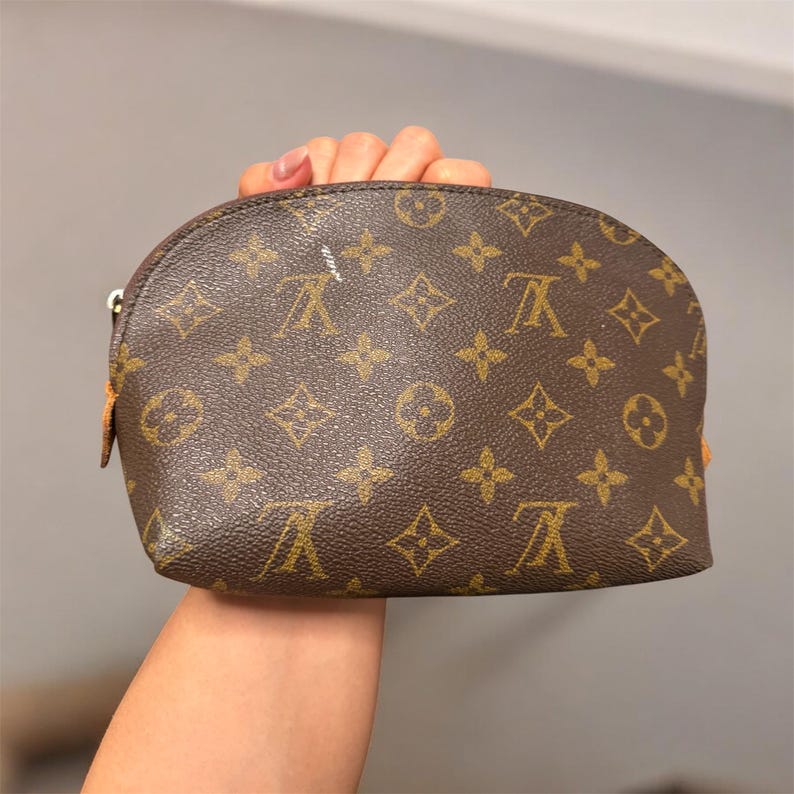 May include: Brown cosmetic bag with a repeating pattern of interlocking letters and floral designs. The bag has a curved top and a zipper closure. The bag is held in a person's hand.