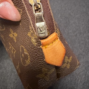 May include: Close-up of a brown and tan monogram canvas pouch with a gold-tone zipper and leather pull tab. The zipper pull features a silver-colored metal with the brand's logo. The pouch is a small, rectangular shape.